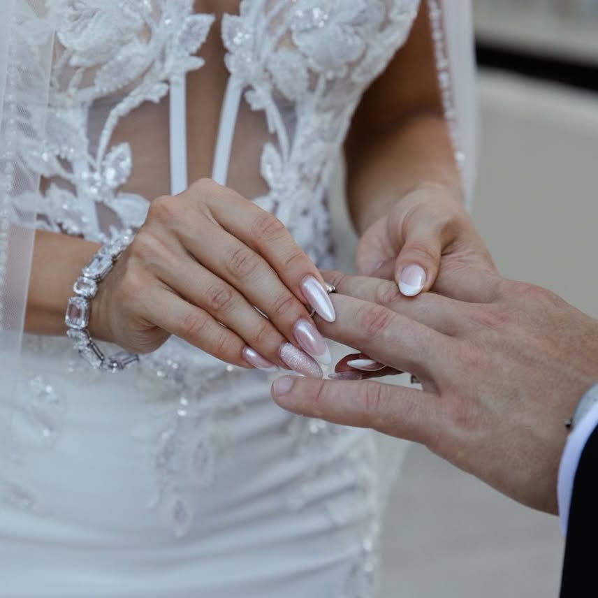 Pearlescent pink bridal manicure with french tips and a glitter accent during a wedding ring exchange